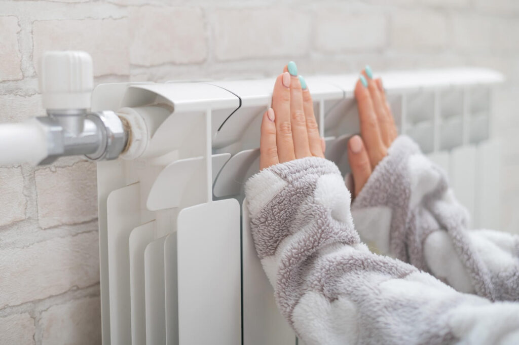 Hands warming over a white radiator heater attached to a light brick‑style bathroom wall